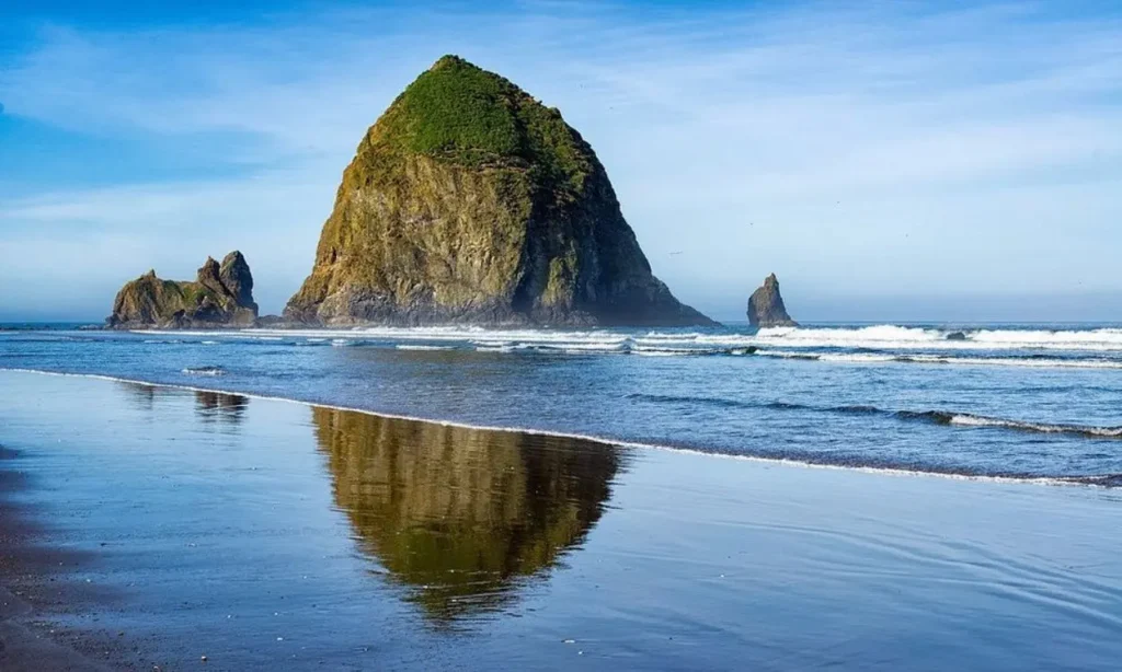 Cannon Beach, Oregon, features Haystack Rock rising from the shore and reflecting on the wet sand.