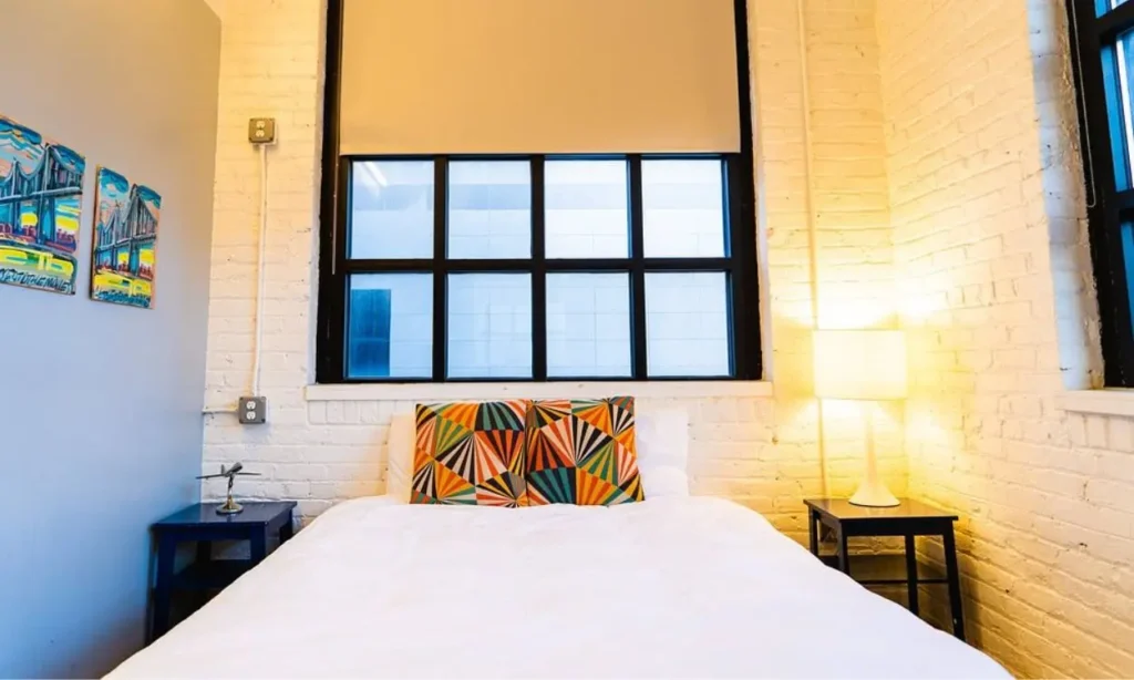 A bright bedroom at The Local NY with a white bed, colorful pillows, and large black-framed windows.
