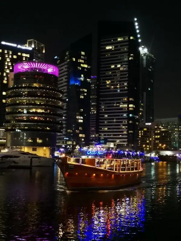 The Alexandra Dhow cruise boat lit up on the water with city buildings behind it.