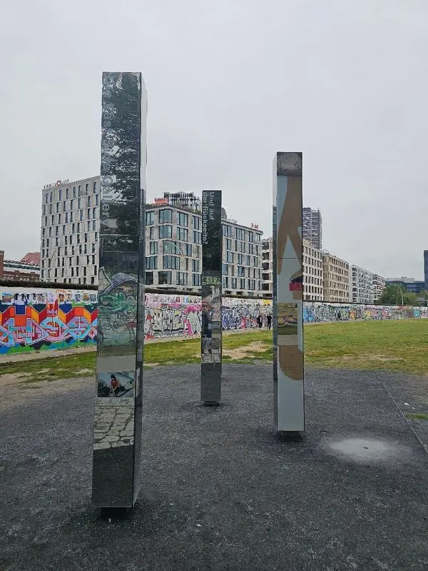 Tall mirrored pillars standing in a grassy area near graffiti-covered walls in Berlin, Germany.