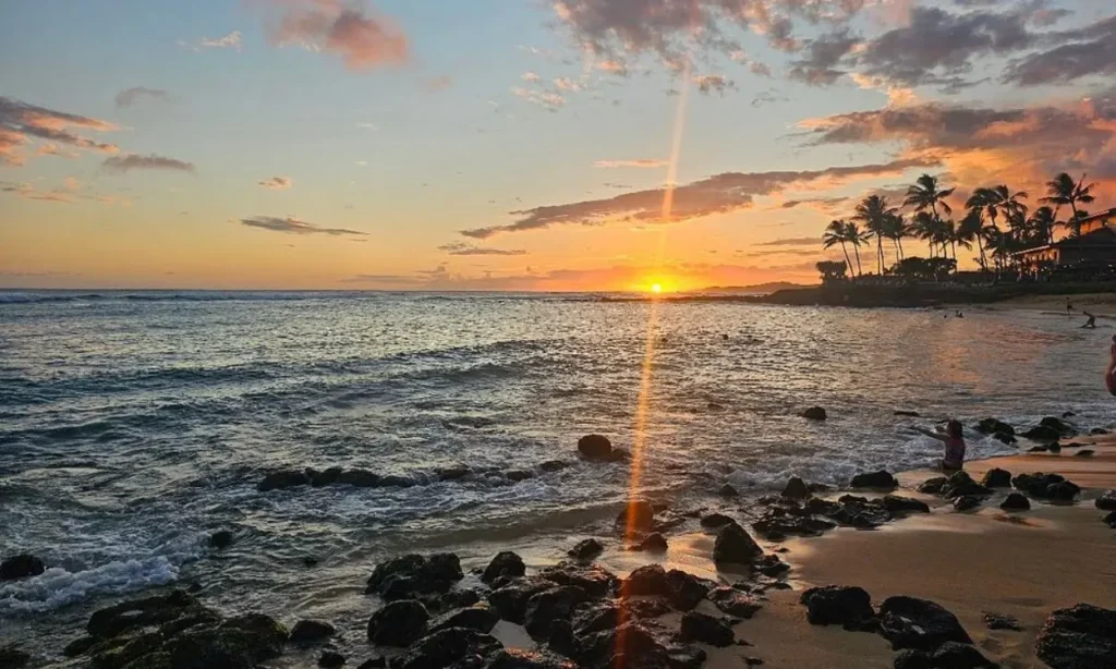 The sun sets over the rocky shore of Poipu Beach, Kauai, Hawaii, with palm trees in the distance.