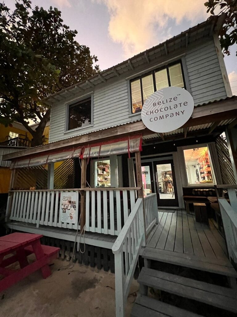 Belize Chocolate Company storefront, a two-story wooden building with a porch, sign, and view into the chocolate shop at dusk.