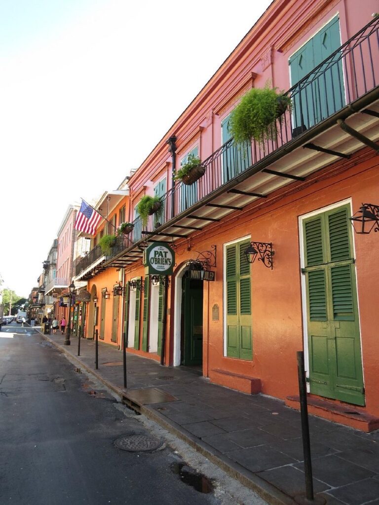 Colorful exterior of Pat O’Brien’s bar in the French Quarter of New Orleans, with green shutters, hanging plants, and an American flag above the street.