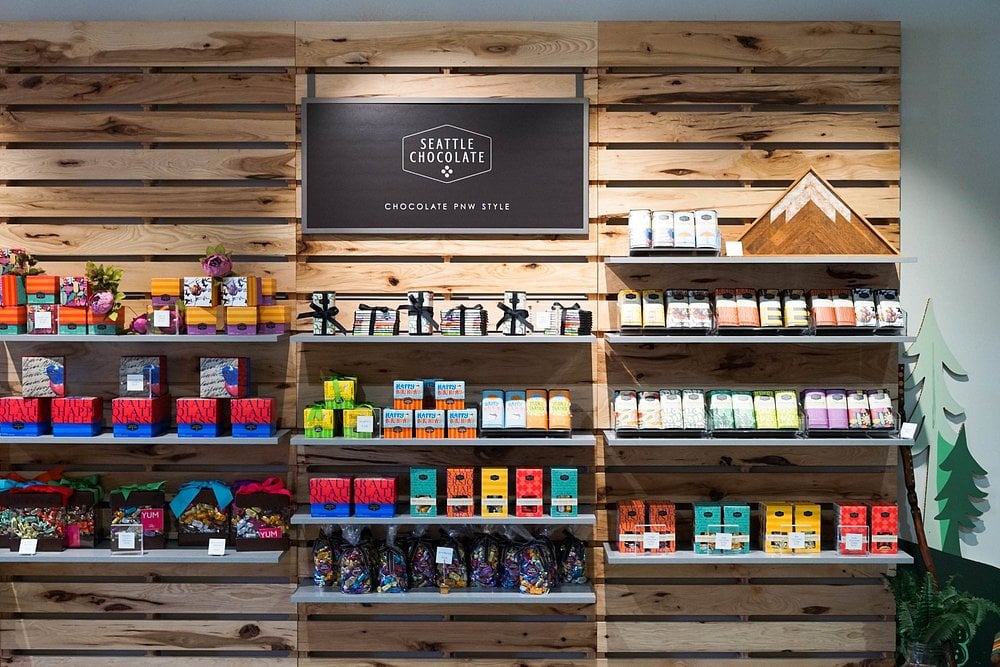 Seattle Chocolate Factory Store display with wooden shelves stocked with colorful chocolate bars, truffle boxes, and gift packages beneath a sign that reads “Seattle Chocolate – Chocolate PNW Style.