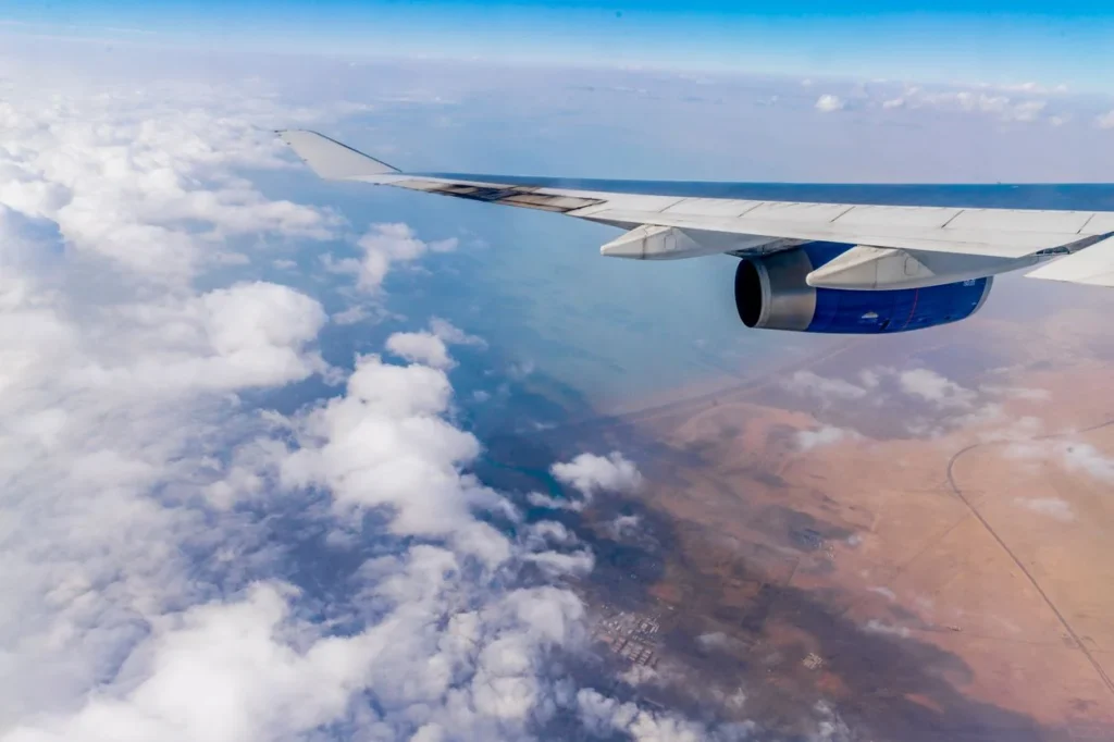 View from an airplane window showing the wing, jet engine, clouds, and a coastline below