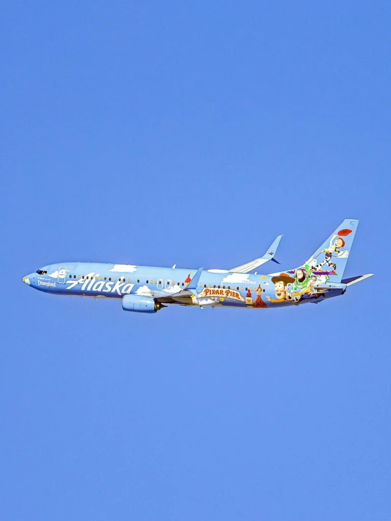 An Alaska Airlines Boeing 737-800 airplane painted in a special blue Disney Pixar Pier livery flying through a clear sky