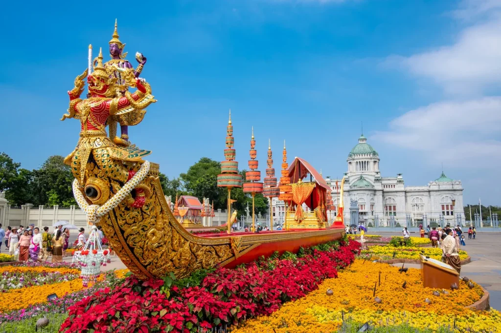 An ornate, golden Thai royal barge replica surrounded by flowers in front of the Ananta Samakhom Throne Hall