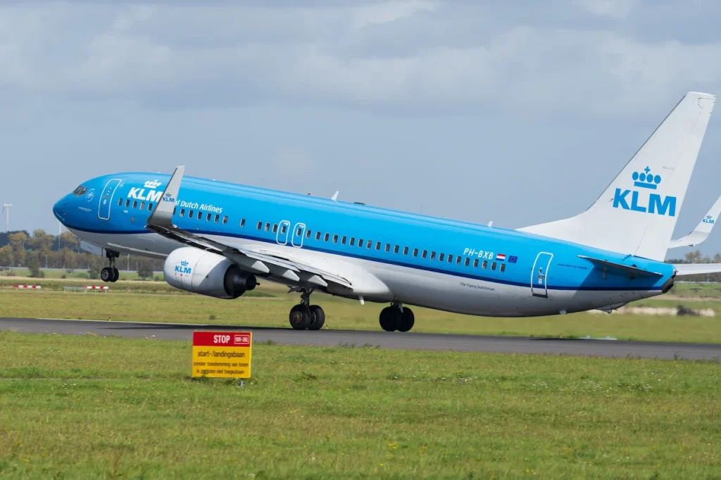 A KLM Boeing 737-800 ascending from an airport runway over green grassy fields