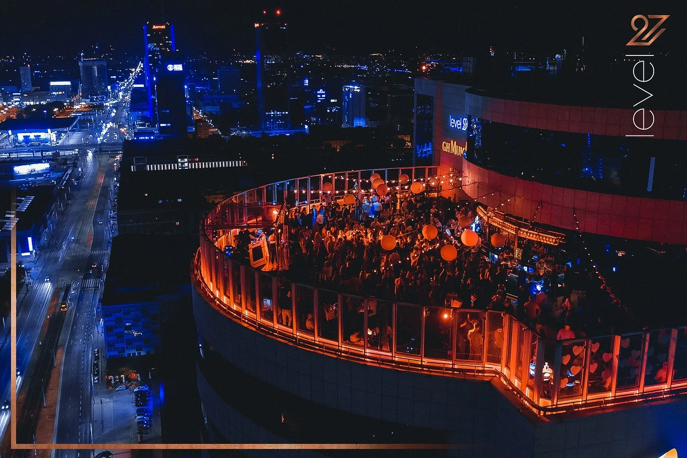 Aerial view of Level 27 rooftop bar terrace at night with crowd of people, orange lighting, and Warsaw city skyline in background