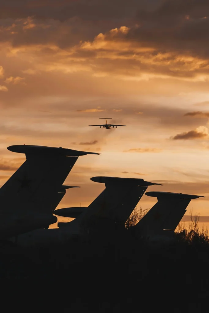 Silhouetted tails of parked military transport planes under a golden sunset with another aircraft airborne