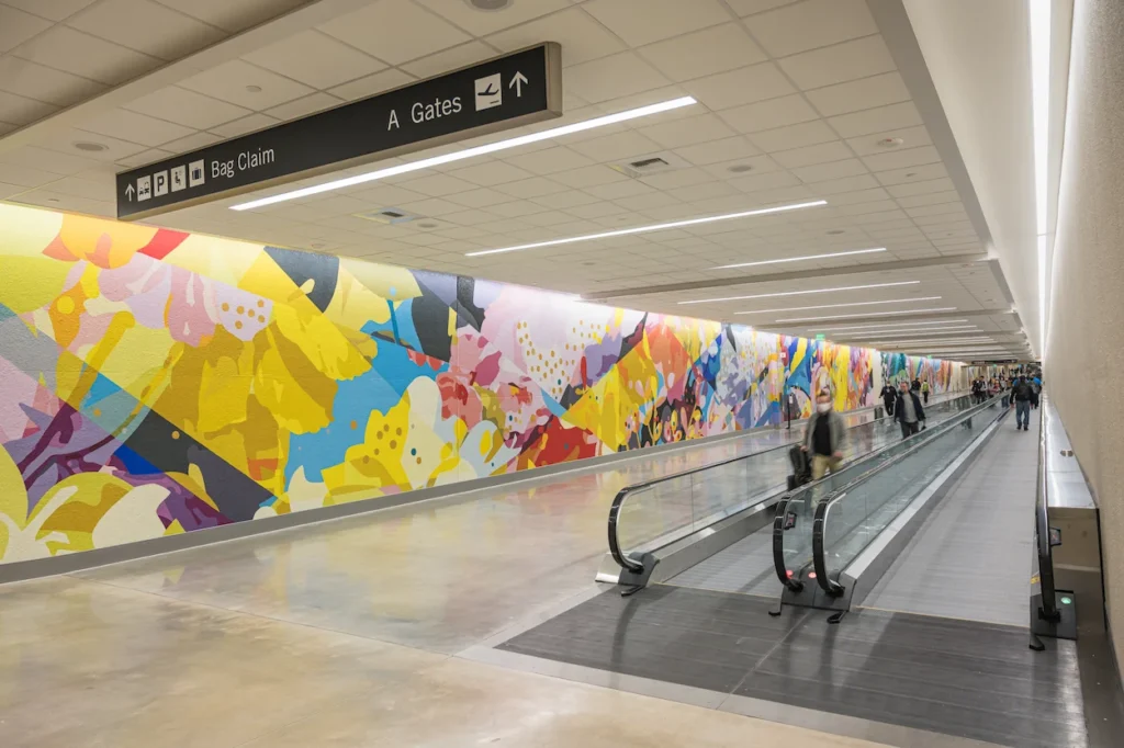 Passengers on a moving walkway alongside a vibrant floral mural inside an SLC Airport tunnel corridor