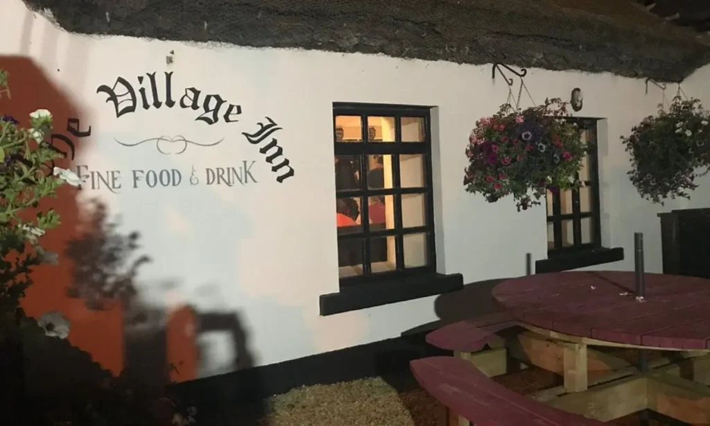 The white-painted exterior of The Village Inn is lit up at night with hanging flowers.