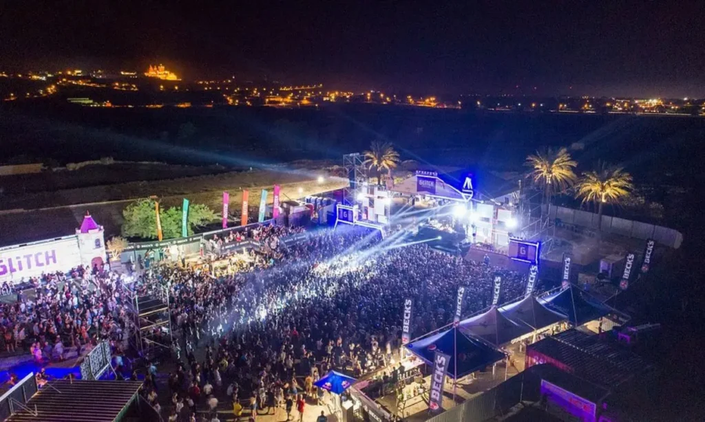 A large nighttime crowd enjoys a music festival at Gianpula Village with bright stage lights.