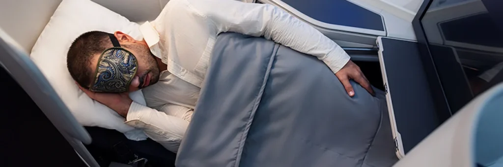 A man sleeping under a gray blanket in an airplane lie-flat bed while wearing a decorative eye mask