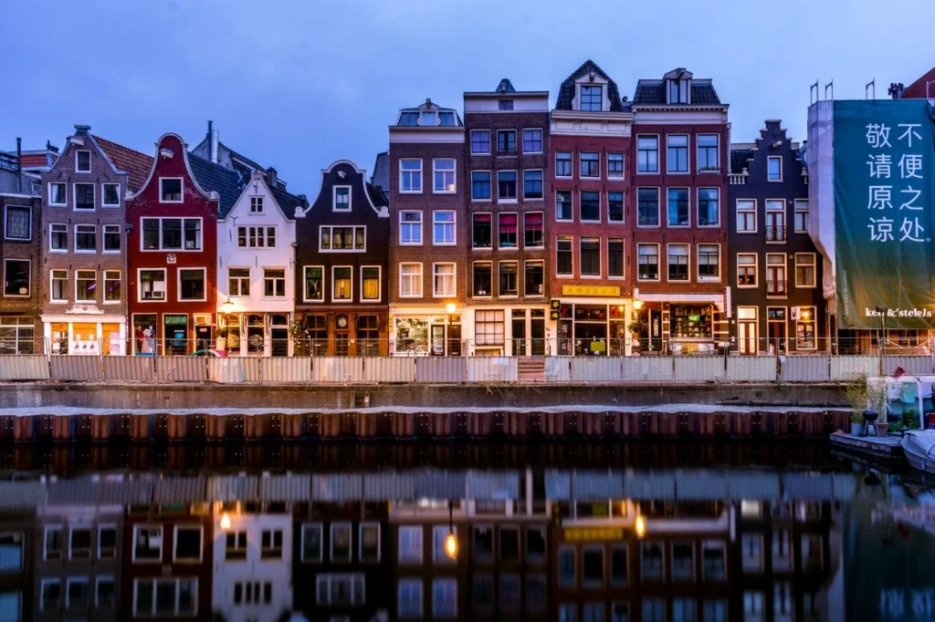 Illuminated narrow Amsterdam houses reflecting on calm canal water at dusk