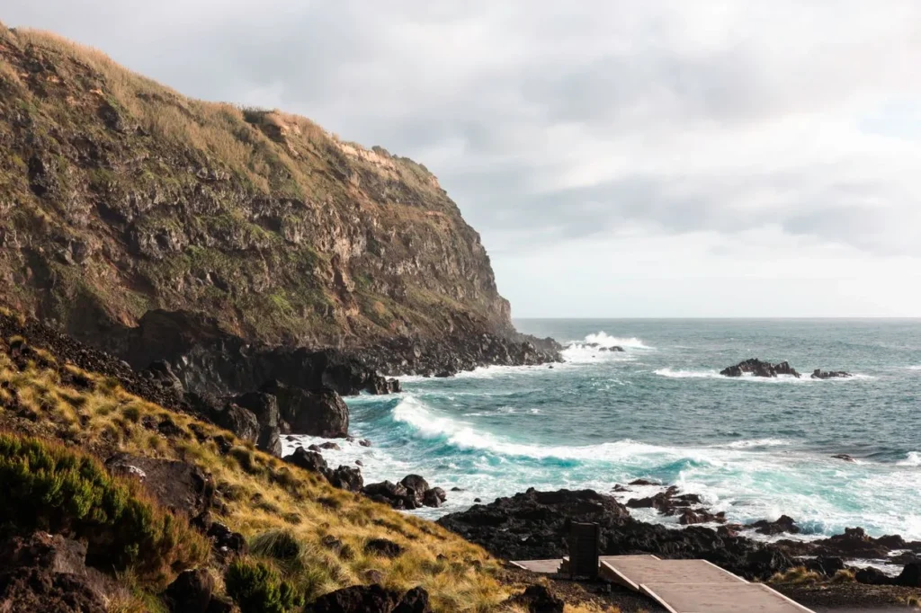 Ocean waves crashing against a rugged rocky coastline with steep, grassy volcanic cliffs under a cloudy sky