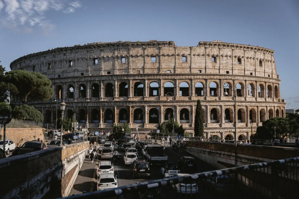 The ancient stone facade of a large amphitheater rising above a busy street filled with cars and people