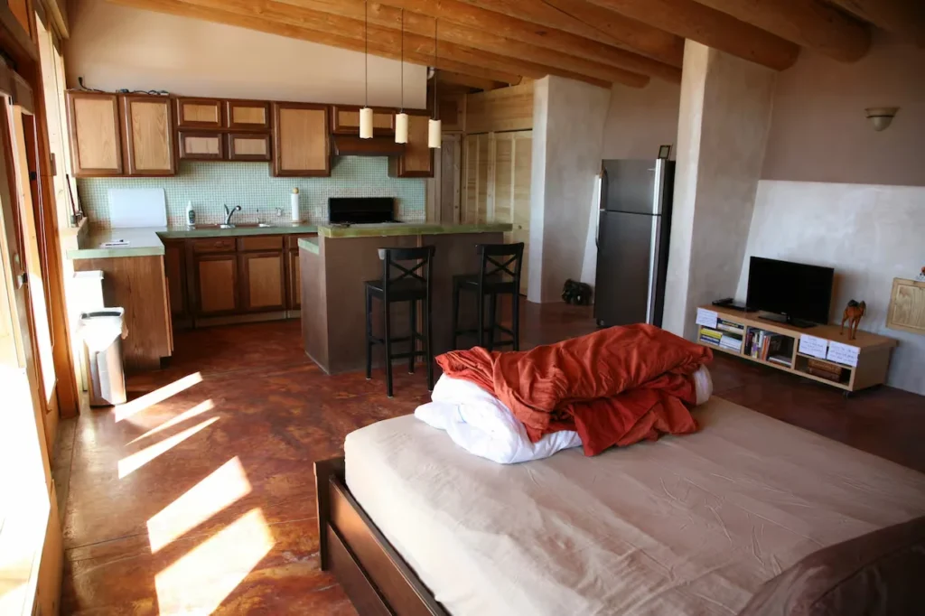 Earthship studio apartment interior with a kitchen bar, stained concrete floor, and a bed in the foreground