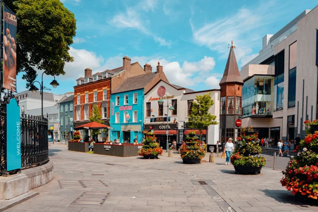 A pedestrian plaza with colorful storefronts and large flower planters at Emmet Place in Cork
