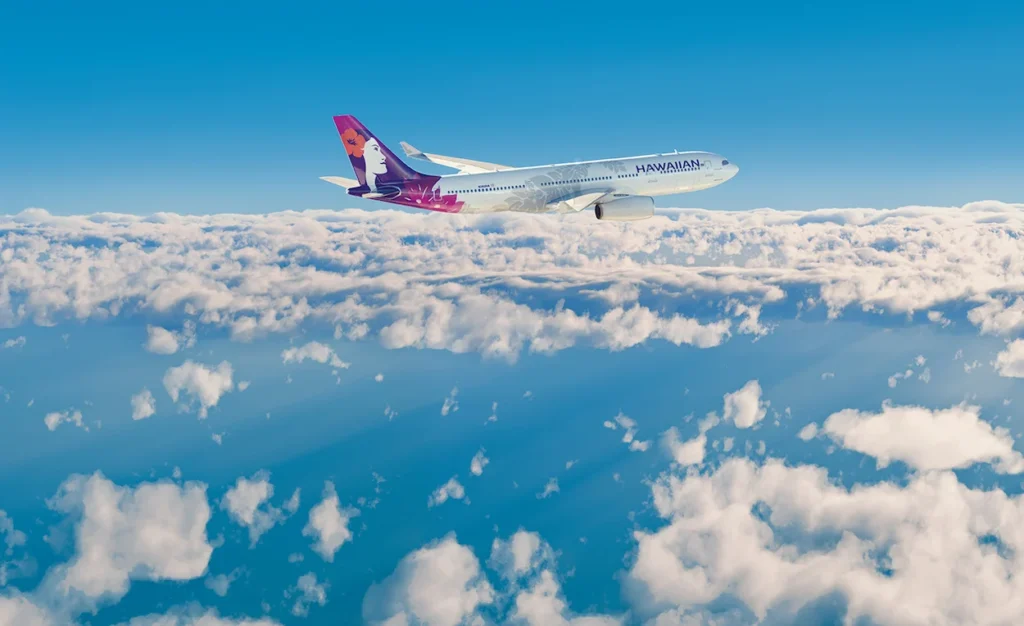 Hawaiian Airlines airplane flying high in the blue sky above a layer of fluffy white clouds