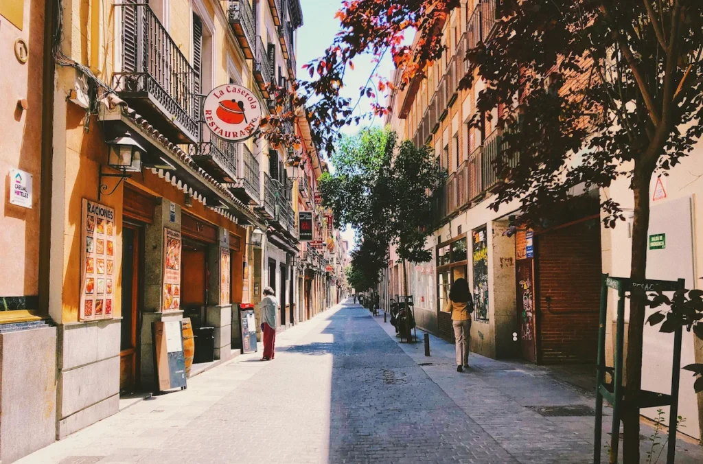 A sunlit narrow street in Madrid featuring the La Caserola restaurant storefront and pedestrians