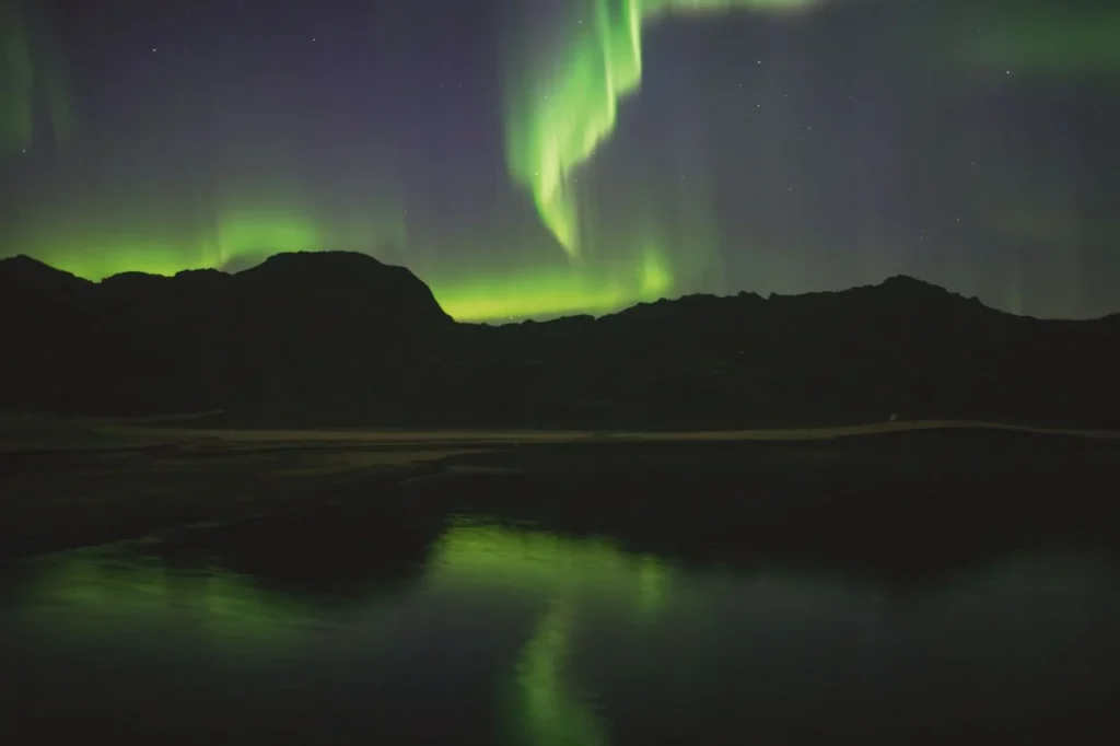 Green northern lights in a starry night sky reflecting on a calm lake behind dark mountains