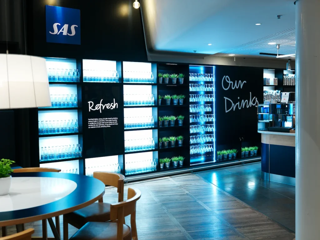 An airport lounge drink station with illuminated shelves of bottled water under an SAS logo sign