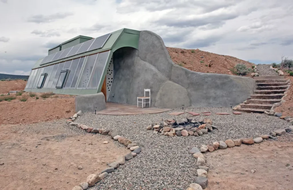 Earthship home built into a dirt hillside with glass windows, solar panels, and a stone fire pit
