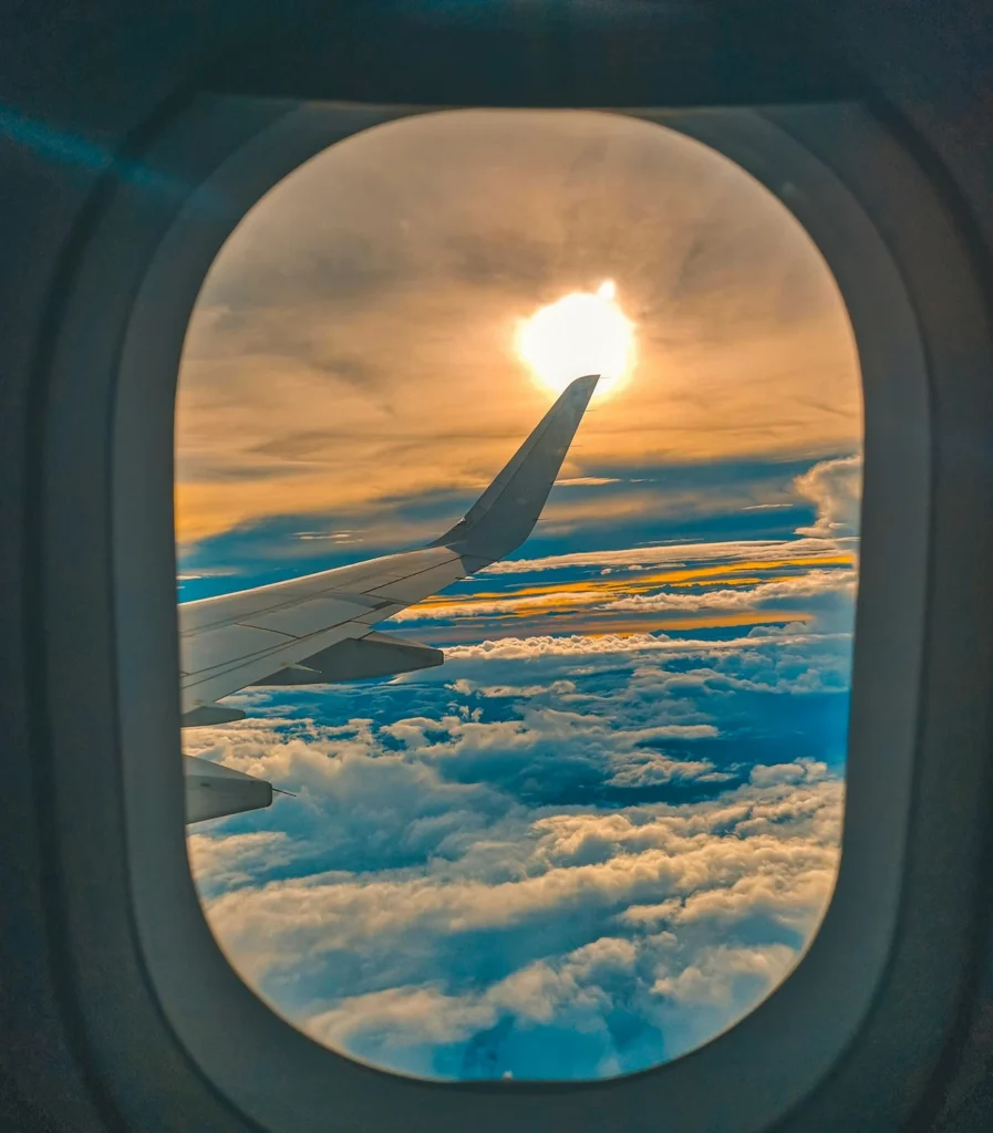View through an airplane window showing the wing, a sea of clouds, and the bright sun
