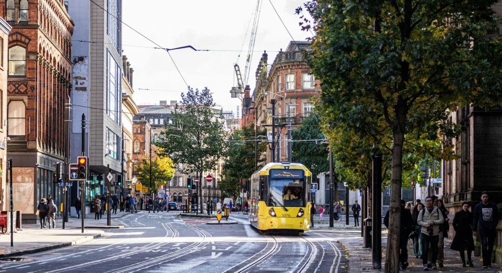 A yellow Manchester Metrolink tram traveling down a city street lined with historic architecture and pedestrians
