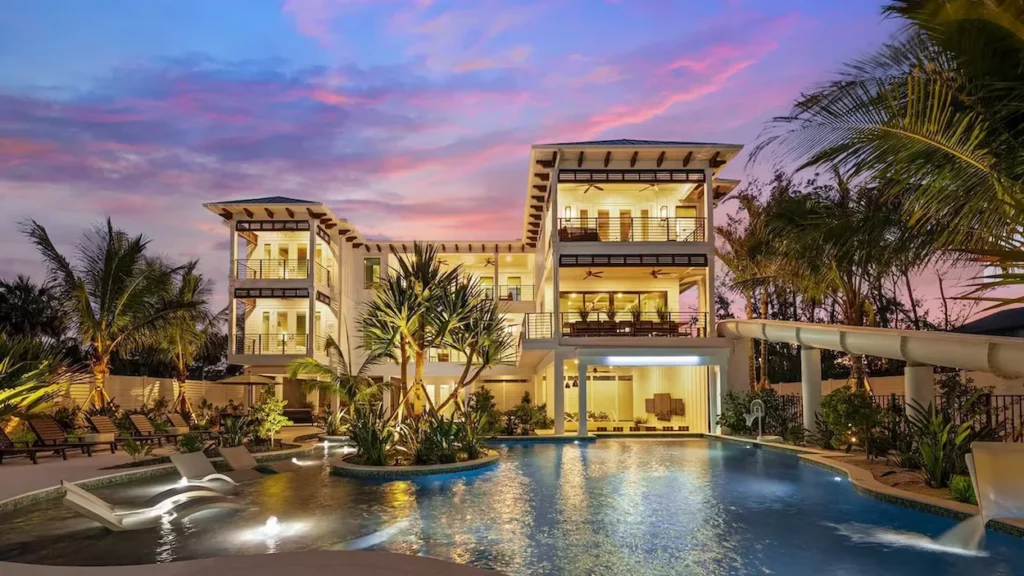 Large multi-story white luxury villa lit up at dusk with a custom swimming pool, palm trees, and an integrated water slide