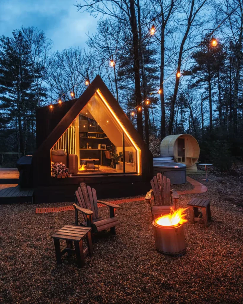 Modern A-frame cabin lit up at night with an outdoor fire pit and barrel sauna