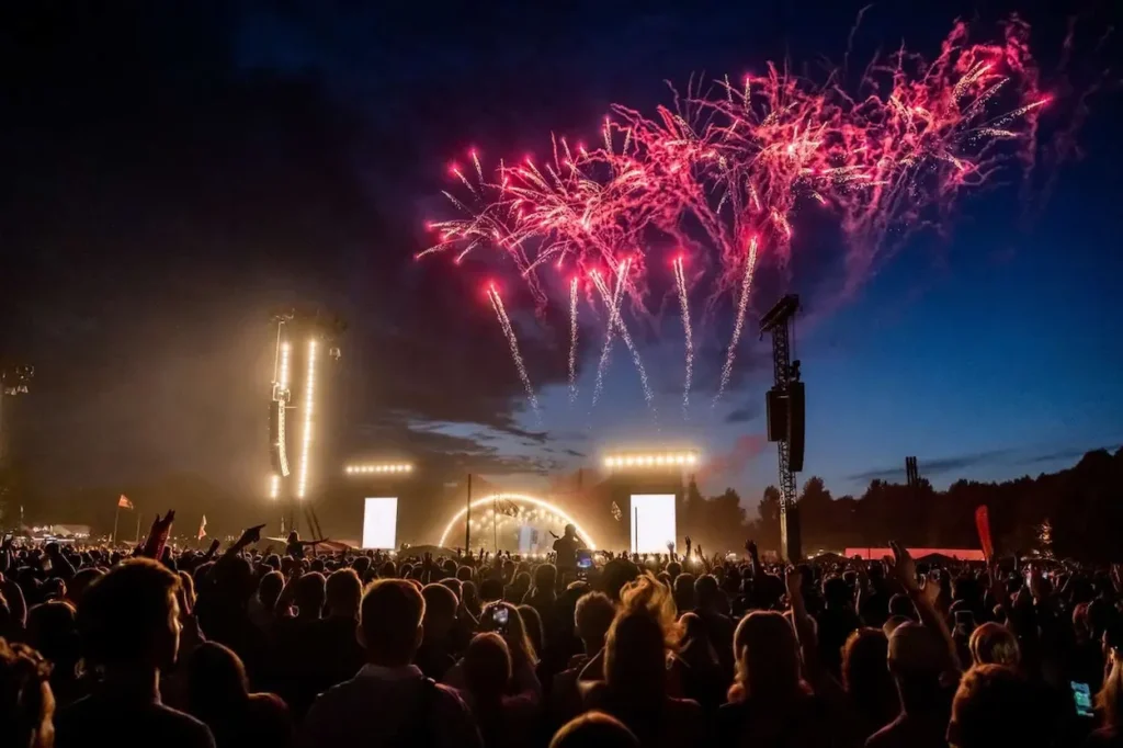 Red fireworks lighting up the night sky above a crowded outdoor concert at the Roskilde Festival Orange Stage