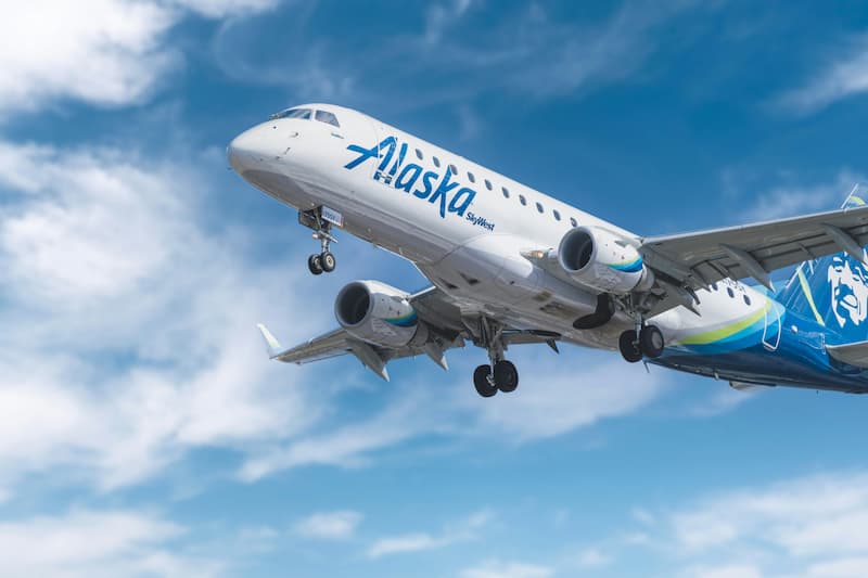 An Alaska Airlines plane takes off into a blue sky with white clouds.