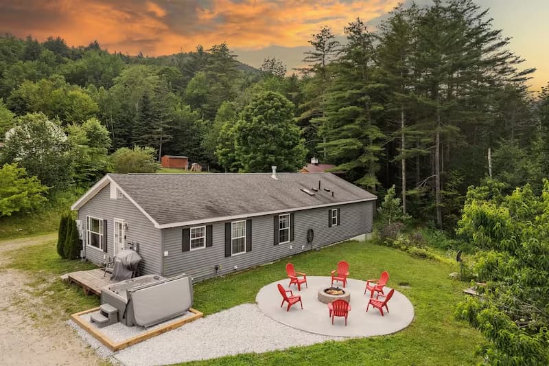 A modern gray house with a hot tub and a circular fire pit area with red chairs.