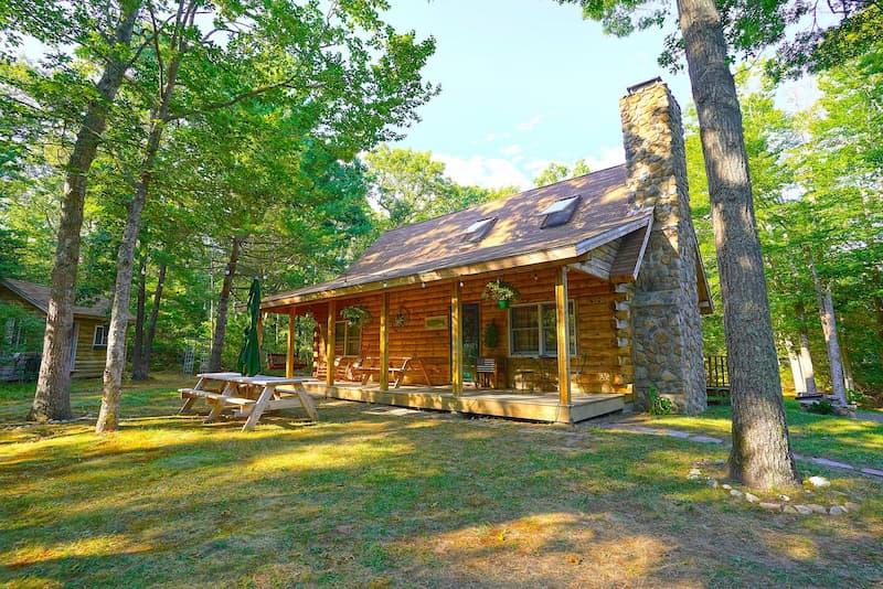 A rustic wooden cabin with a stone chimney, surrounded by green trees.