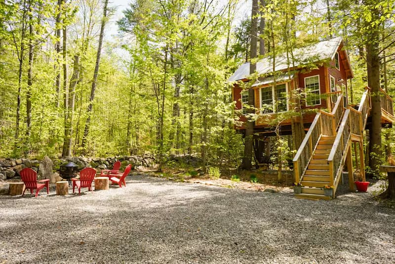A red treehouse with a staircase leading up, surrounded by green trees and a fire pit area.