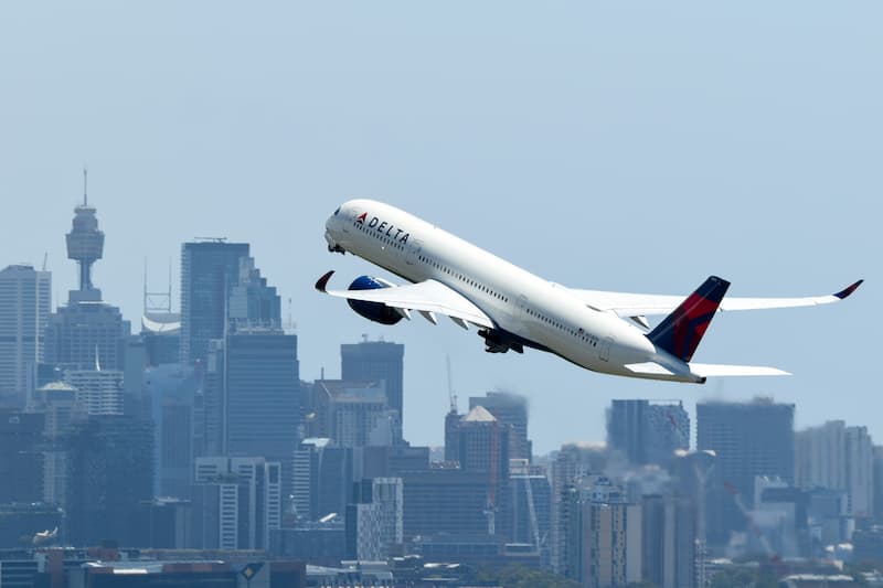 A Delta Air Lines plane takes off with a city skyline in the background.