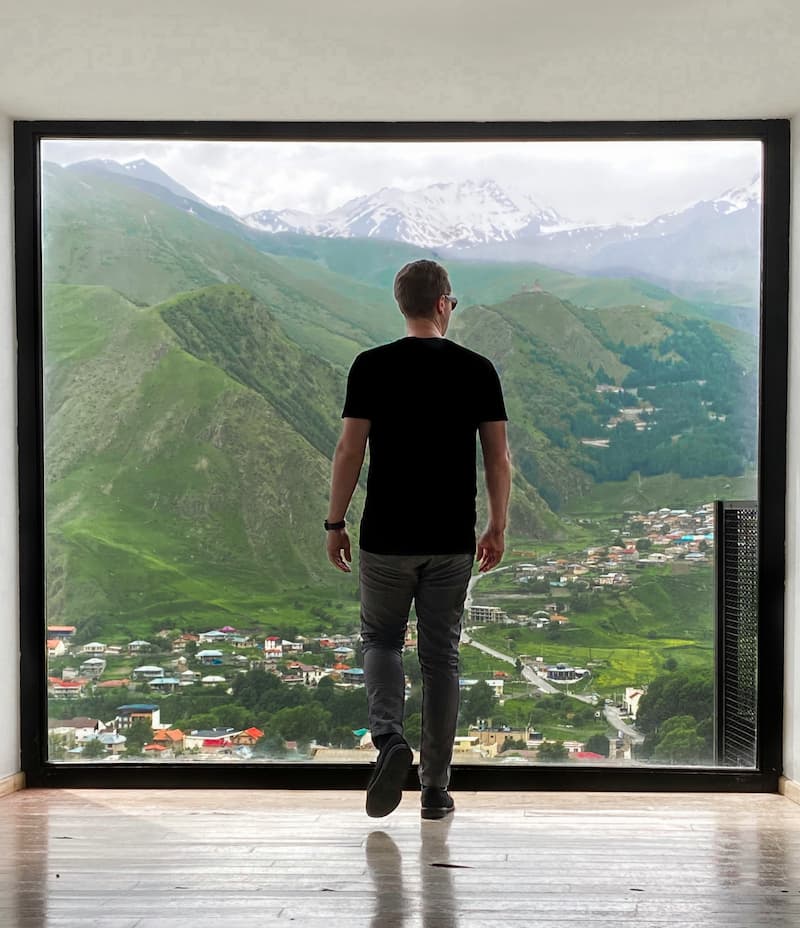 A person stands by a window, overlooking a town nestled in a valley surrounded by mountains.