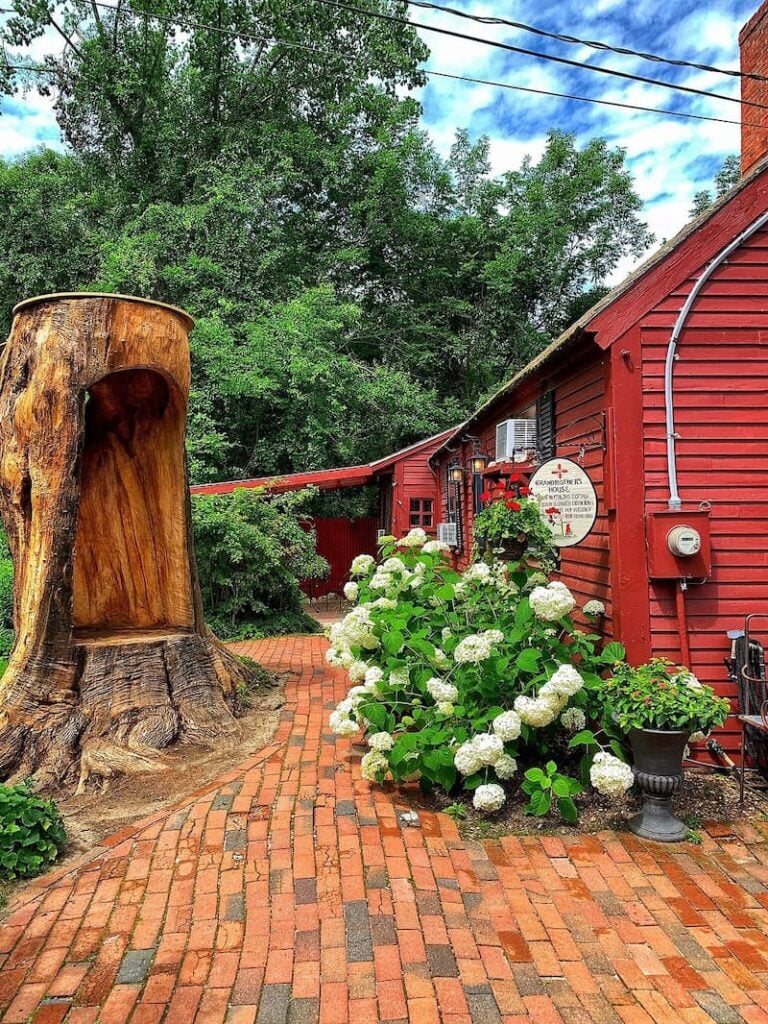 A red cottage with a brick patio, hydrangea bush, and a large, hollowed-out tree stump.