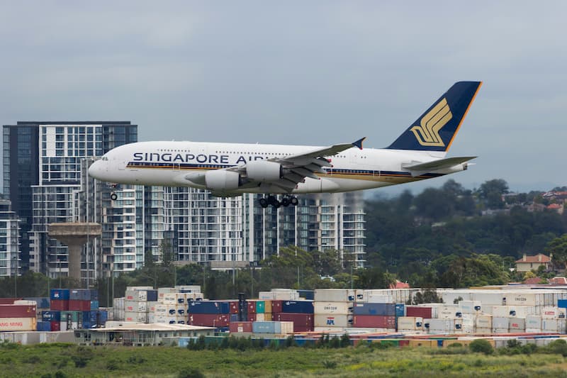 A Singapore Airlines plane descends over a city with buildings and shipping containers.