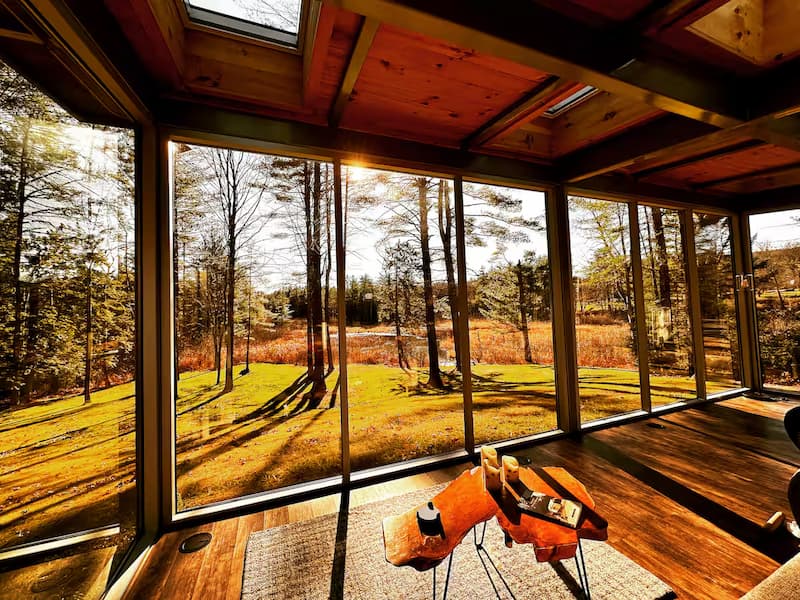 Interior view of a glass house overlooking a sunlit forest and field.