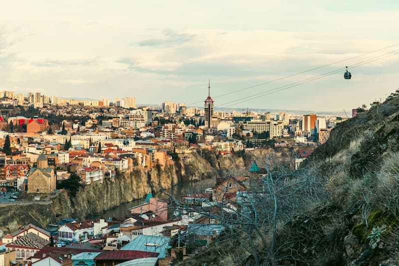 Panoramic view of Tbilisi, Georgia, with old city buildings, a river, and distant skyline.