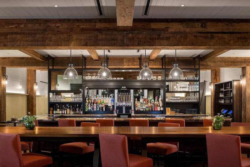 A well-lit bar area with wooden beams, a long communal table, and modern pendant lights.