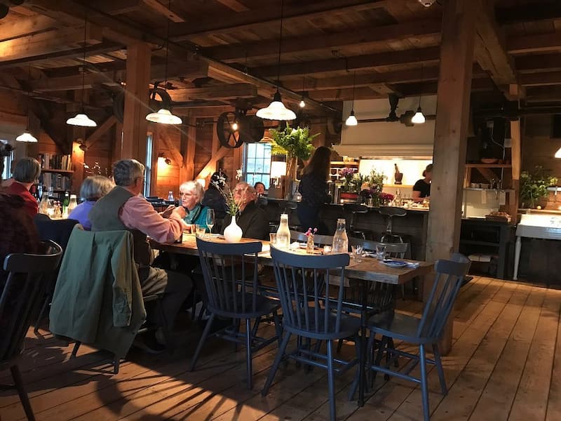 The rustic, warmly lit interior of a barn restaurant with diners at long wooden tables.
