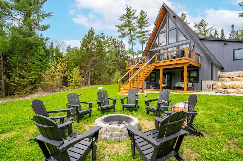 An A-frame house with a large deck and a fire pit surrounded by chairs on a green lawn.