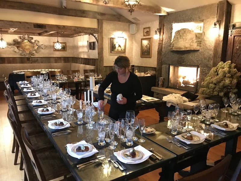 A staff member sets a long, elegant dining table in a rustic pub with a stone fireplace.