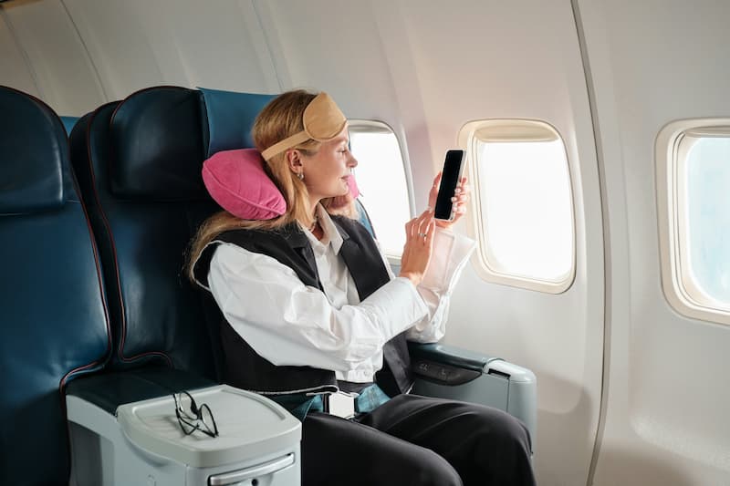 A woman with an eye mask and neck pillow using a smartphone next to an airplane window.