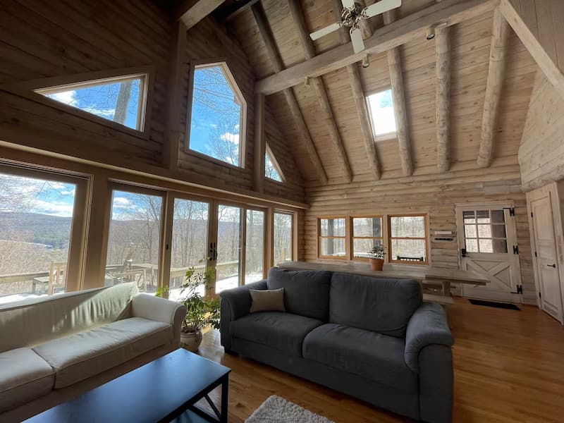 Interior of a log cabin with large windows offering a snowy mountain view, and a living area.