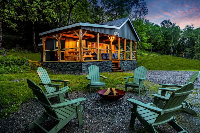 An illuminated cabin with a screened porch at dusk, and a fire pit with green chairs.